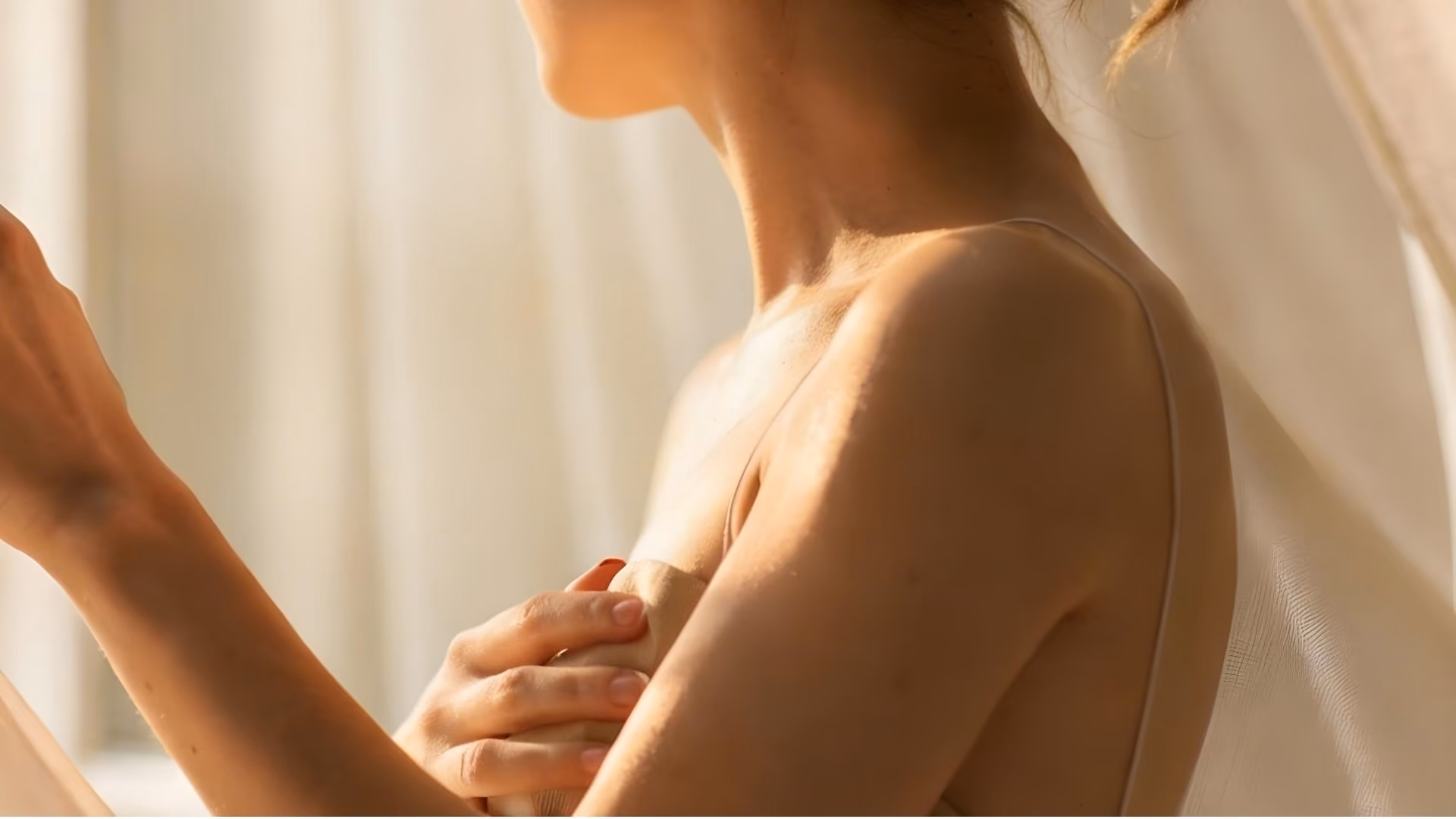 A woman in soft window light beside a sheer curtain, seen from the shoulder, one hand resting at her chest.
